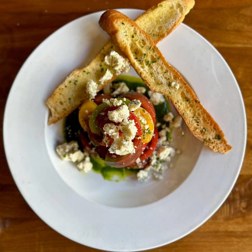 Heirloom tomato salad with crumbled feta cheese, green sauce, and two garlic bread slices on a white plate.