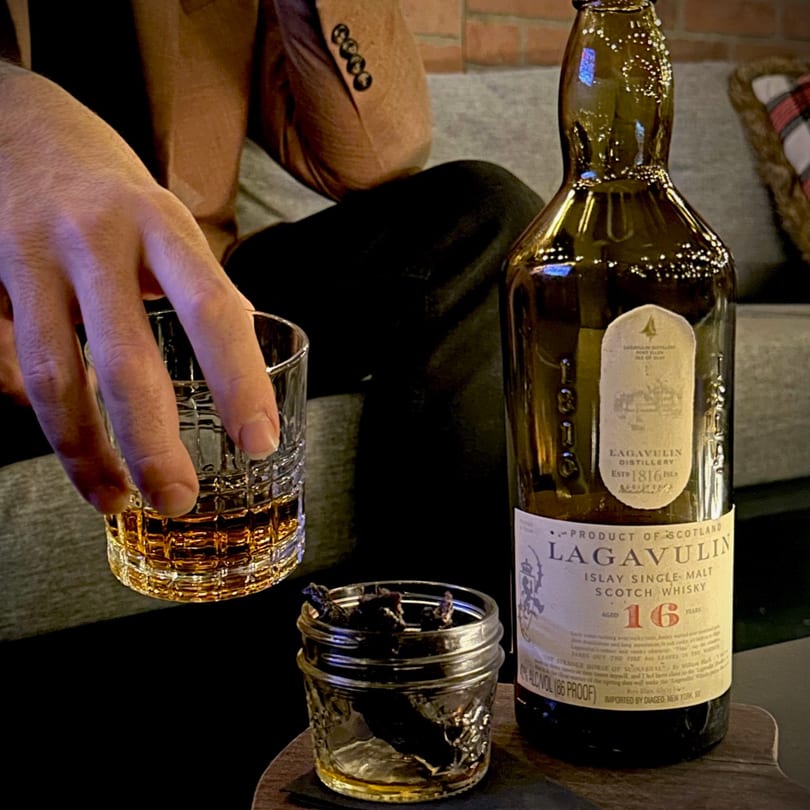 A person holds a glass of amber whiskey next to a bottle of Lagavulin and a jar of jerky.