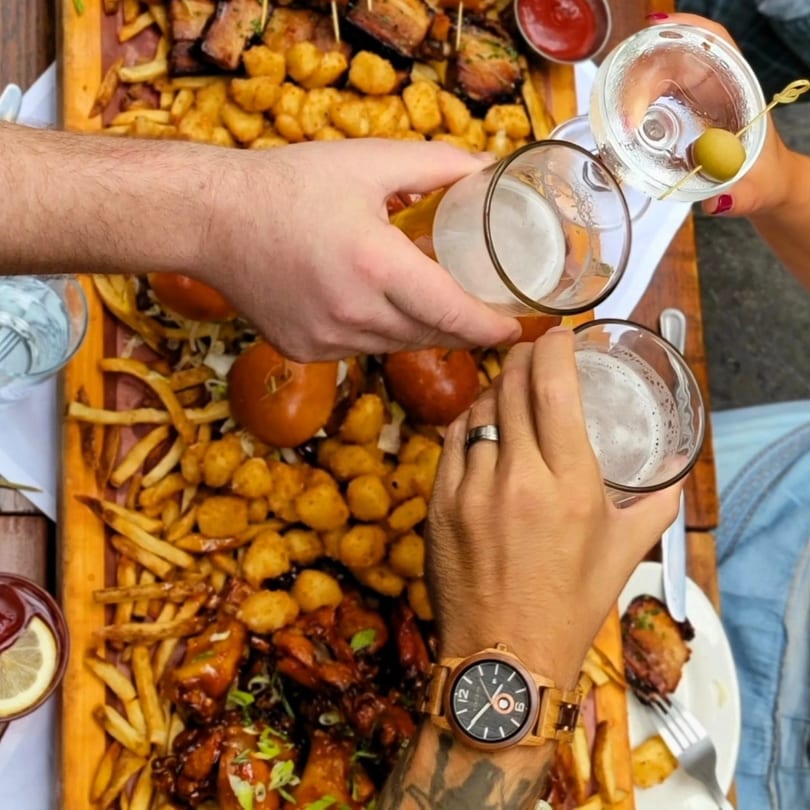 Four people toast drinks over a large Butcher Bar platter of fries, tots, wings, and sliders.