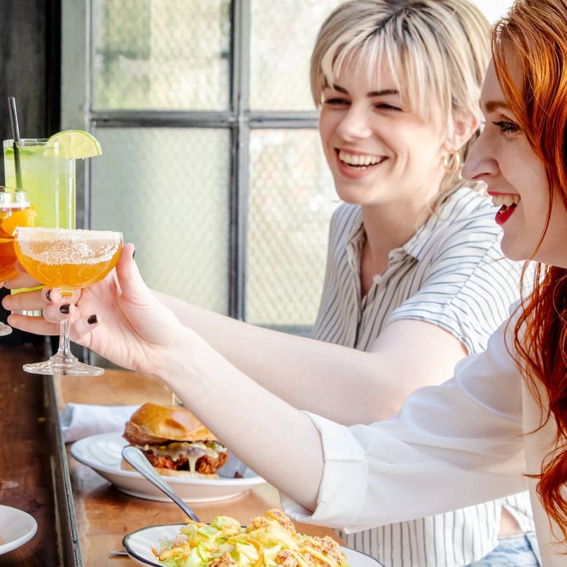 Two women laughing and toasting cocktails at a bar-and-grill, with food on the table.