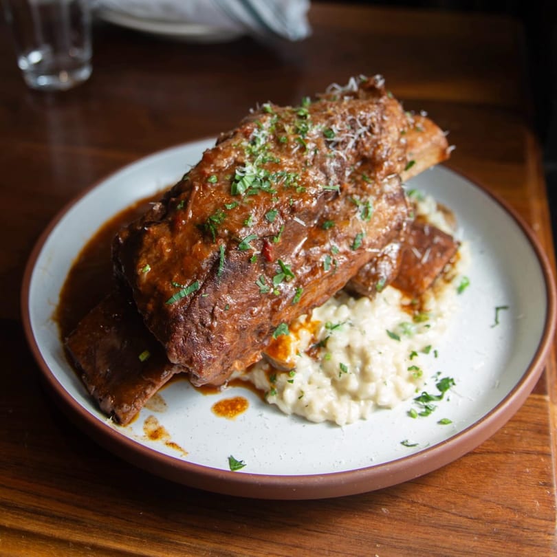 Braised short ribs with gravy and risotto, garnished with parsley and parmesan, on a white plate.